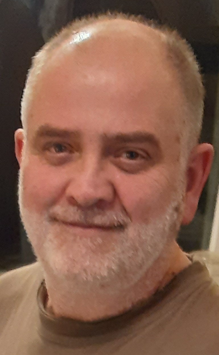 Muž s šedým plnovousem a plešatou hlavou, opírá bradu o prsty před knihovnou.